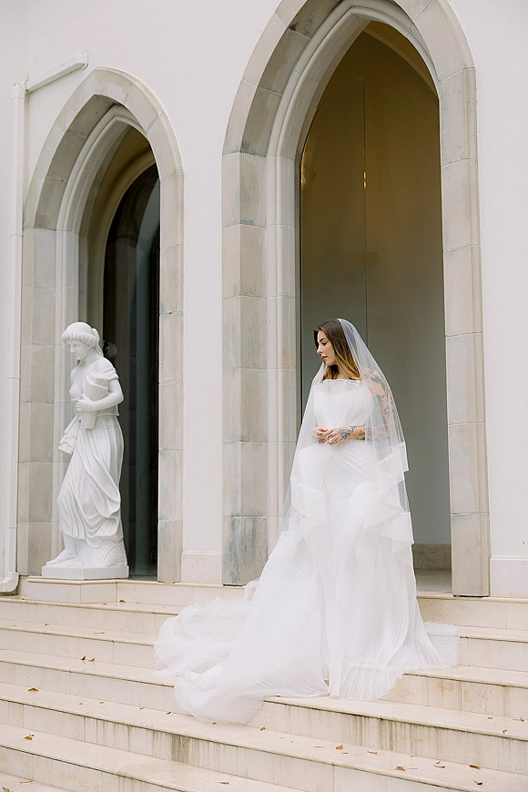 Bridal portrait of a bride on steps, looking down in a strapless wedding dress with long train and veil at an arched doorway
