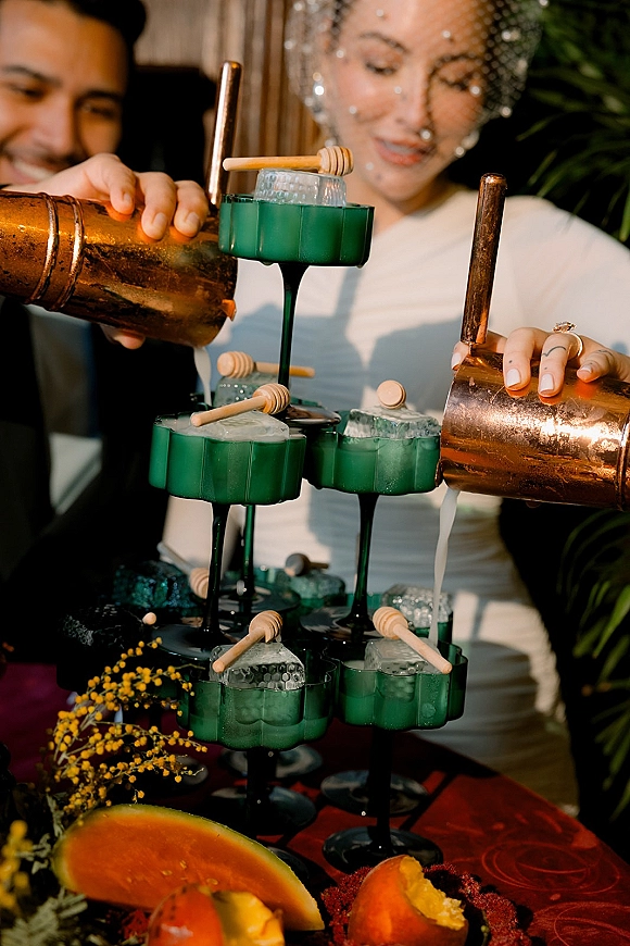 Champagne tower pour cascading through green coupe glasses, with bride’s birdcage veil and ring visible at an indoor bar setup backdrop