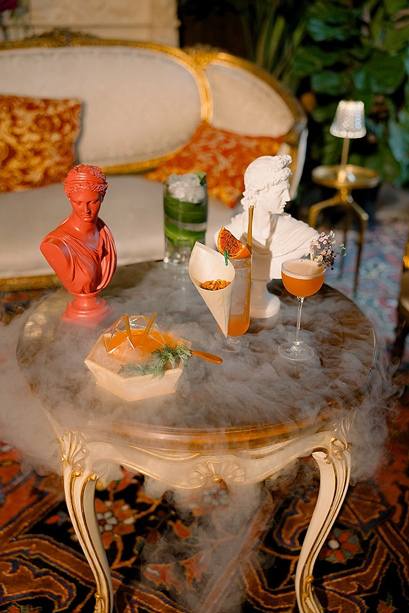 Wedding cocktail table with signature wedding cocktails, citrus garnish, ice bucket, and appetizer cones on a marble coffee table in an indoor lounge