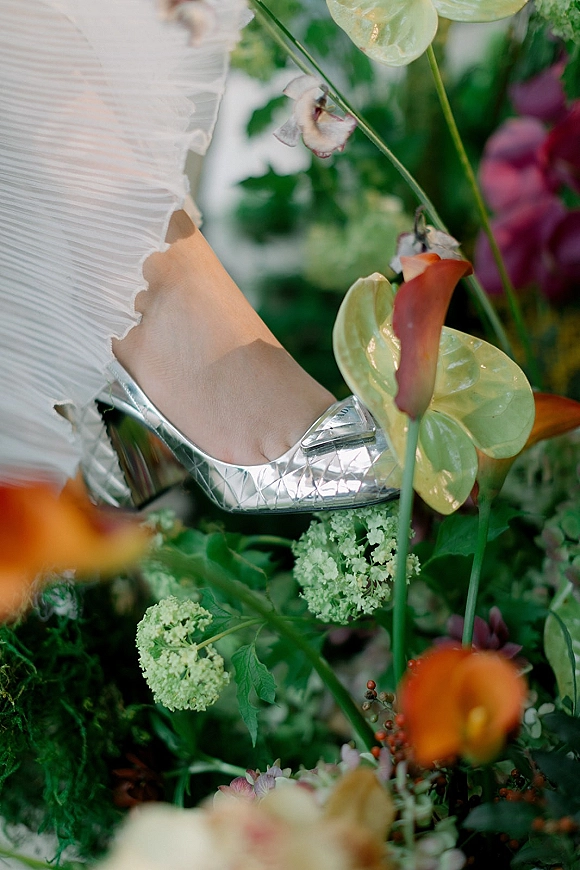 Bridal shoes, silver wedding shoes nestled in garden florals beside a pleated dress hem with anthurium and calla lilies accents