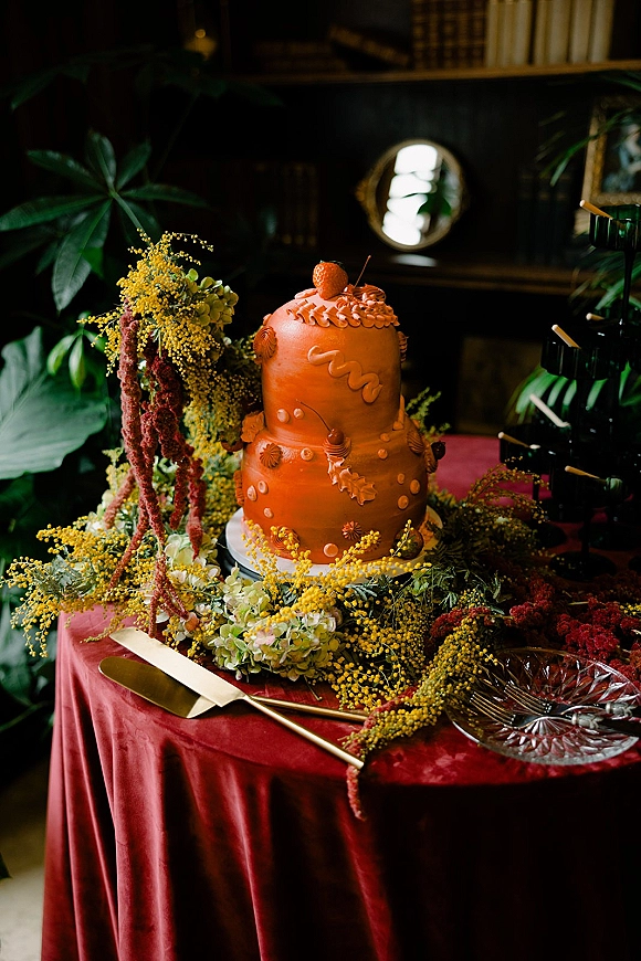 Wedding cake with strawberry topper, a two-tier buttercream design on a red velvet table with hydrangeas and black taper candles in a moody library setting