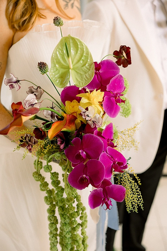 Bridal bouquet with tropical bridal bouquet orchids, anthurium, and calla lilies, with trailing greenery against a bride’s gown indoors
