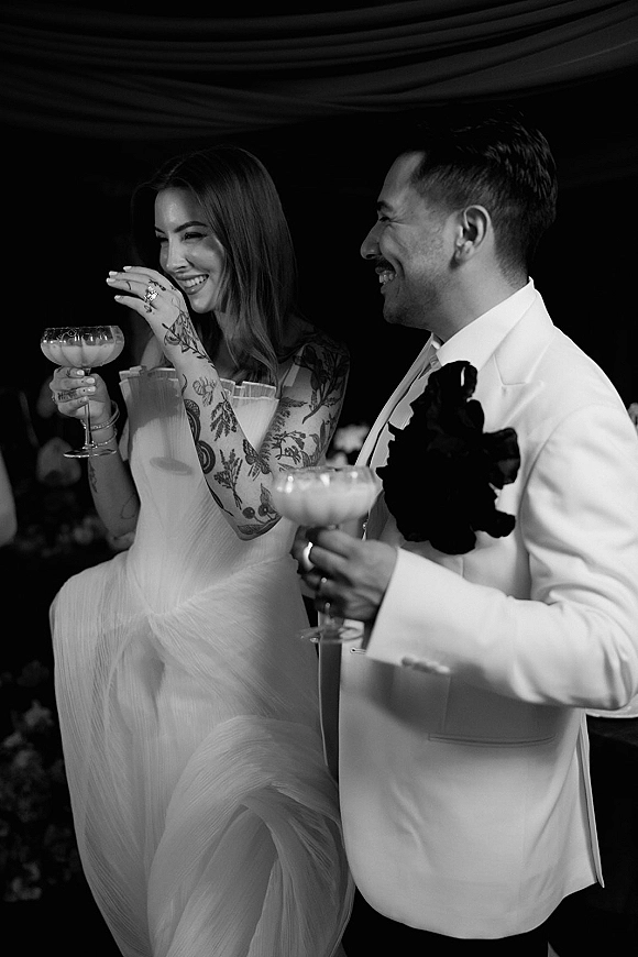 Reception toast as bride in strapless dress laughs with groom in white tuxedo jacket, raising coupe glasses under draped ceiling in dark space