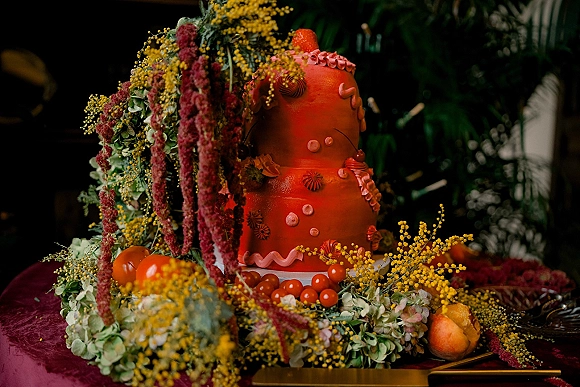 Wedding cake with red frosting, piped icing, and strawberries, grapes, and citrus on a velvet table amid moody greenery backdrop