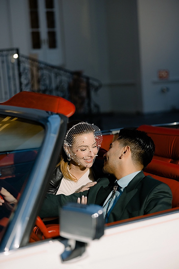 Couple portrait of bride and groom in car, leaning in to kiss in a vintage convertible with red leather seats and birdcage veil accent