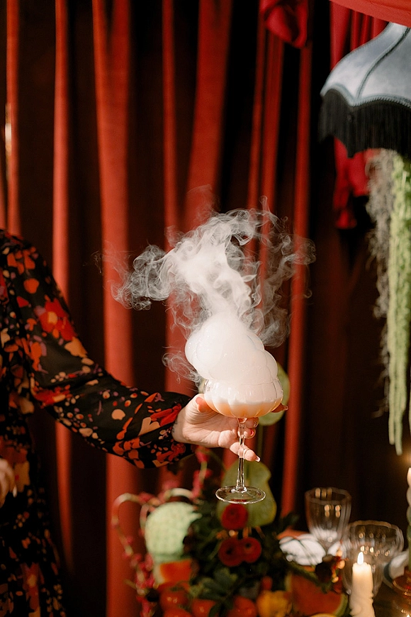 Smoking cocktail with dry ice smoke rising from a foamy coupe glass on a candlelit reception table before red drapery backdrop