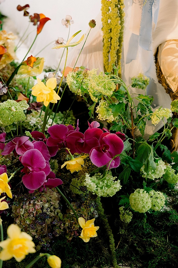 Wedding floral arrangement with orchids and daffodils in a ceremony floral meadow, featuring moss, greenery, and a hanging garland on draped fabric backdrop