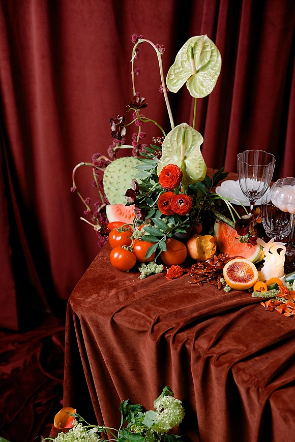 Wedding tablescape with a moody wedding tablescape vibe, anthurium and ranunculus florals, fruit slices, candles, goblets, and a draped backdrop