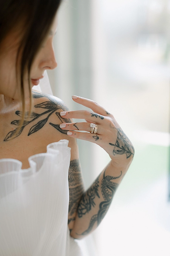 Wedding rings and engagement ring close-up on a tattooed bride’s manicured hand in soft window light with a blurred outdoor view