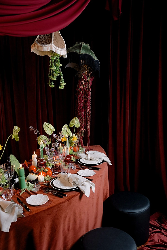 Reception tablescape wedding table setting with taper candles, citrus and florals, vintage lampshades, and velvet curtain backdrop in a dark interior