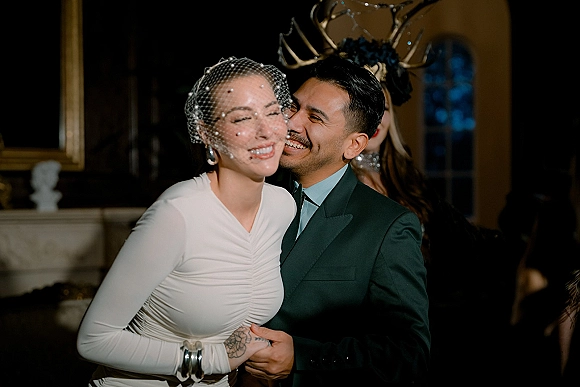 Wedding couple portrait of bride and groom laughing as he kisses her cheek, bride in birdcage veil and long-sleeve dress in dim venue