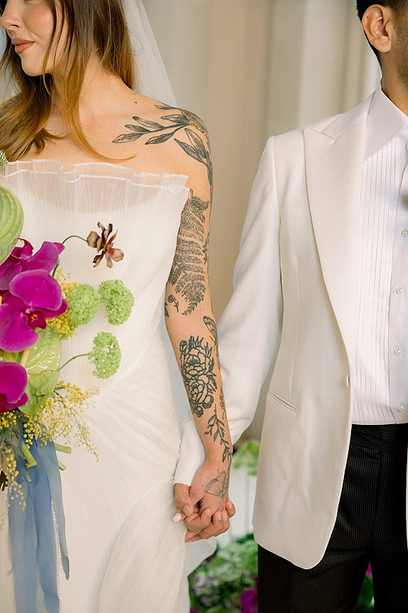 Couple portrait of bride and groom holding hands, showing tattooed bride in strapless gown with veil and colorful bouquet with blue ribbon indoors