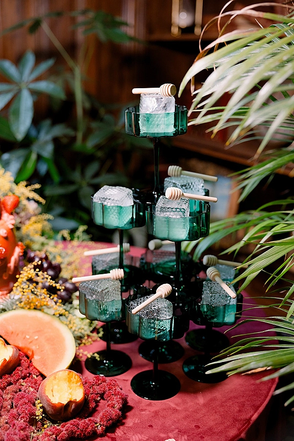 Wedding drink station with green glass goblets, ice cubes, honey dippers, citrus and grapes on a burgundy tablecloth by a wood wall