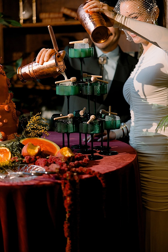Wedding cocktail tower with a tiered cocktail stand of stacked coupe glasses holding emerald green drinks, citrus garnish, and raspberries in warm indoor reception lighting