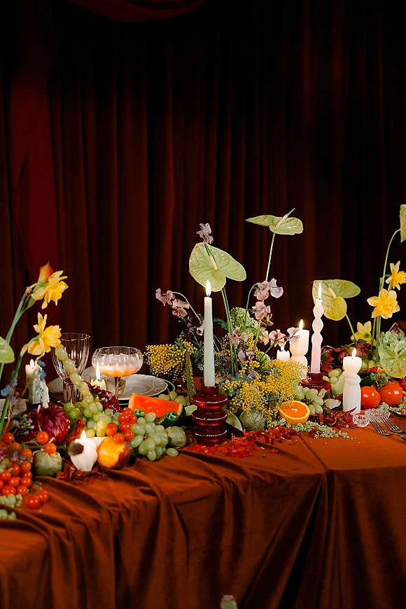 Reception tablescape with colorful wedding tablescape accents, twisted taper candles, fruit and florals on rust linens before a draped backdrop