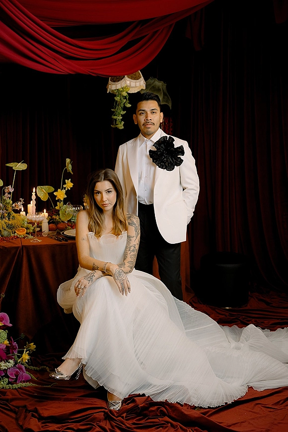 Couple portrait of seated bride in strapless tulle gown and standing groom in white tuxedo with oversized lapel flower at candlelit table, red drapery backdrop