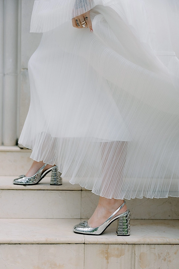 Wedding shoes in silver bridal heels on stone steps beside a pleated tulle skirt, with wedding band and engagement ring on display