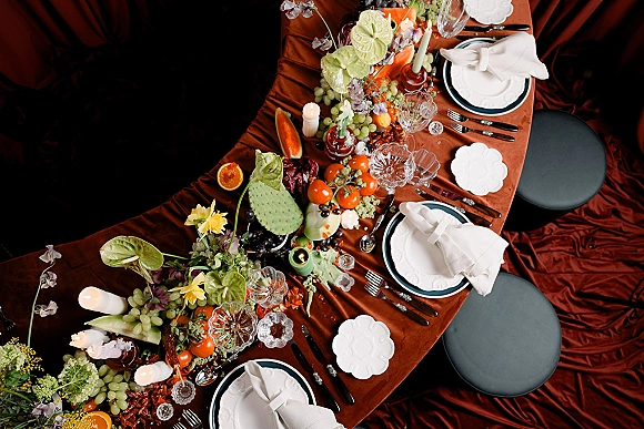 Reception tablescape overhead with rust velvet tablecloth, taper candles, crystal glassware, orchids, and fruit centerpieces on dark floor