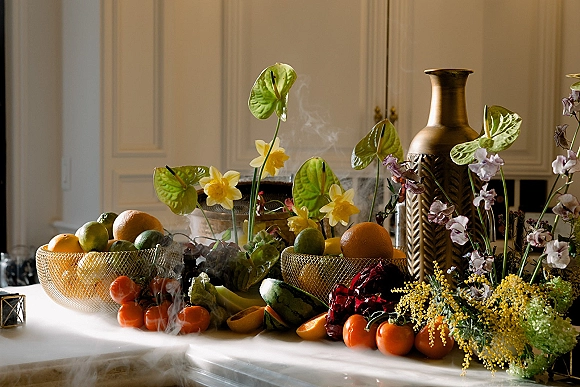 Wedding tablescape with a citrus wedding centerpiece in brass vase, mesh fruit bowls, anthurium and daffodils on a tabletop by a white paneled wall