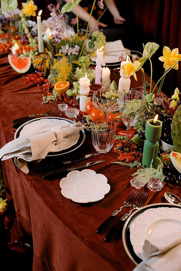 Reception tablescape with wedding table centerpiece of daffodils and greenery, tapered candles, and fruit on a brown tablecloth in a moody interior
