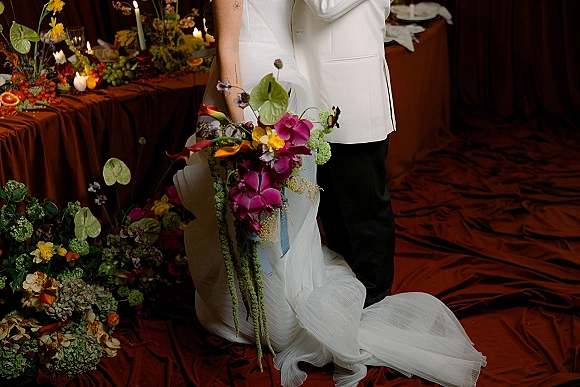 Wedding couple portrait of bride and groom embrace beside a candlelit reception table, bride holding cascading orchid bouquet against draped backdrop