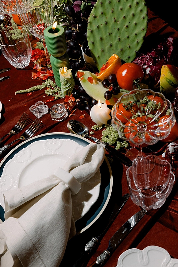 Reception tablescape with wedding place setting on rust tablecloth, green taper candles, crystal votives, and fruit with cactus pad centerpiece