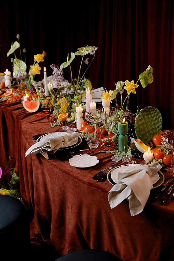 Reception tablescape with velvet tablecloth wedding table, taper and pillar candles, floral greenery garland and fruit centerpiece before dark drapery