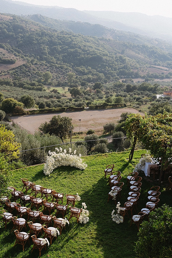 Ceremony setup with outdoor ceremony seating in a curved layout, white floral ceremony arch and aisle arrangements on a vineyard lawn with mountain valley views