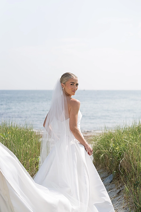 Bridal portrait of a bride looking back in a strapless wedding dress and long veil, with earrings, against ocean and dune grass shoreline