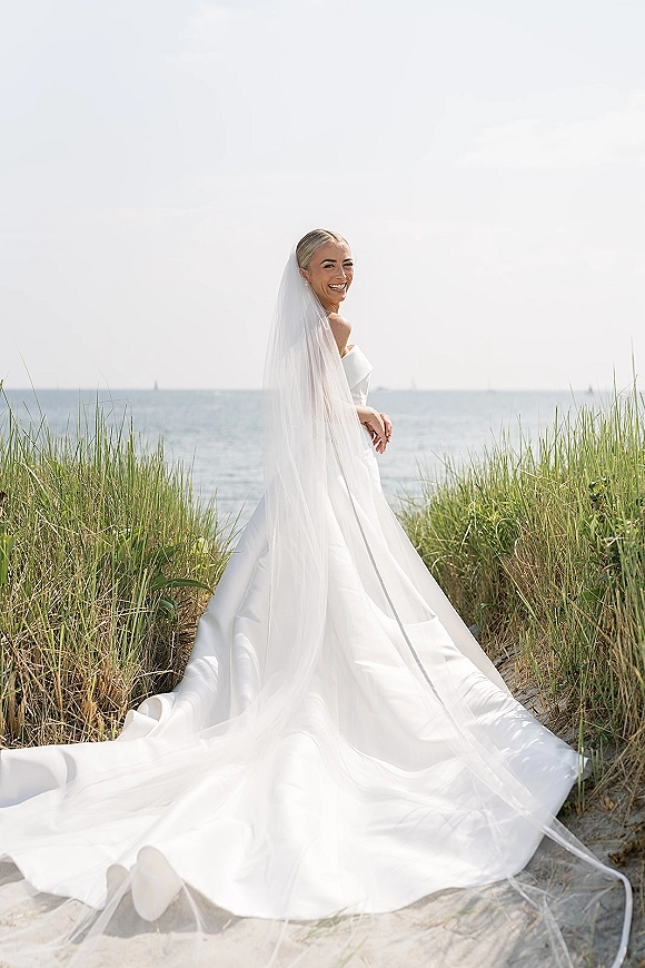 Bridal portrait of a beach bride in a wedding dress with long veil and train, standing in dunes with tall grass, ocean, and sky behind