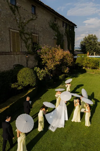 Processional moment as bride walks with bridesmaids, one holding a white parasol, across manicured lawn toward groom by ivy-clad villa