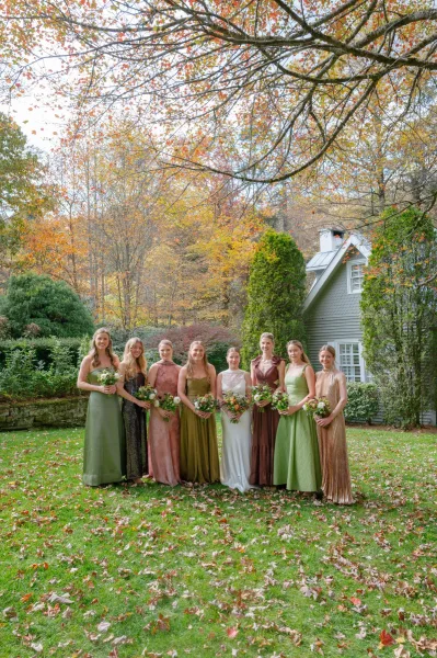 Bridesmaid group photo with bride and bridesmaids holding bouquets in mixed dresses on a garden lawn with autumn trees and stone wall