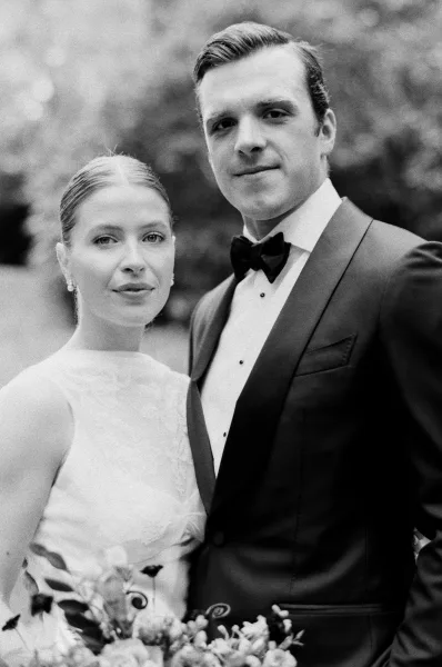 Couple portrait in black and white, bride in lace high-neck dress holding bouquet beside groom in tuxedo with bow tie amid trees