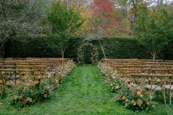 Ceremony setup with outdoor wedding ceremony seating, wood crossback chairs facing a floral arch with roses and greenery on a fall lawn