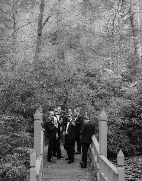 Groomsmen portrait of men in tuxedos wearing sunglasses and raising beer cans on a wooden footbridge over pond water in the forest