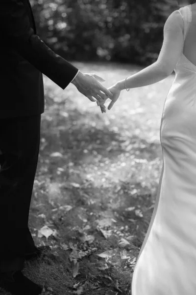 Couple holding hands as they walk, bride in a sleek wedding dress with a bracelet beside groom in a suit amid leafy greenery