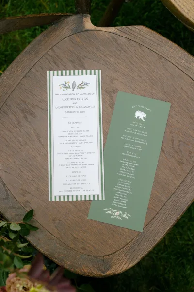 Wedding programs with striped border and botanical illustration on green cardstock, arranged on a wood table beside garden greenery