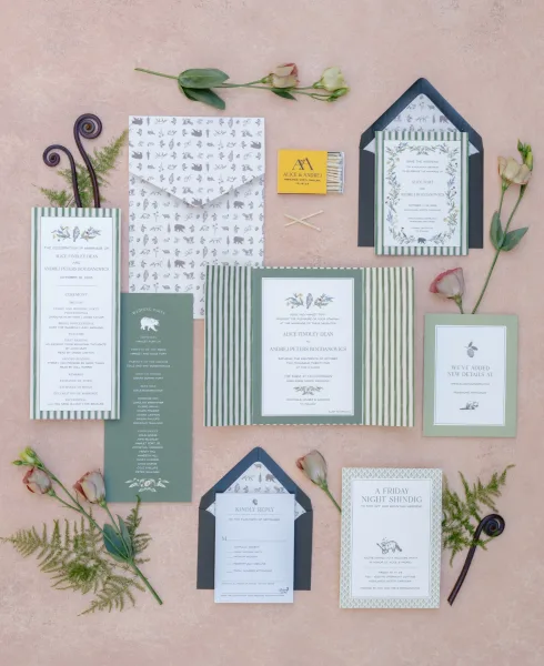 Invitation suite wedding invitation flat lay with green striped cards, navy envelopes, wax seals, matchbox, roses, and greenery on blush surface