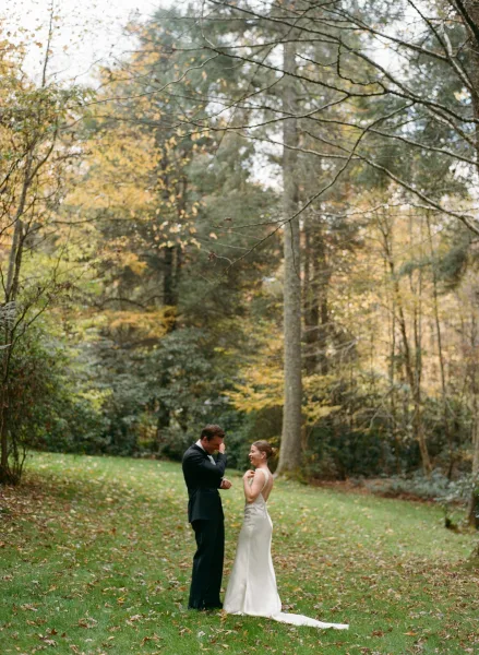 First look moment as bride in a low back gown with train faces groom in suit and boutonniere on a fall forest lawn with leaves