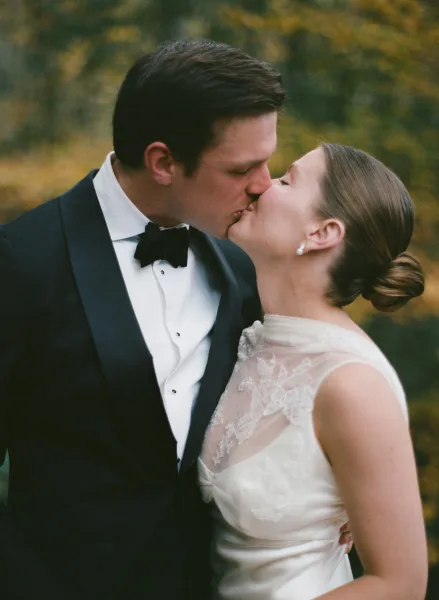 Wedding kiss portrait of bride and groom kissing, groom in black tuxedo and bow tie, bride in lace bodice gown amid autumn trees