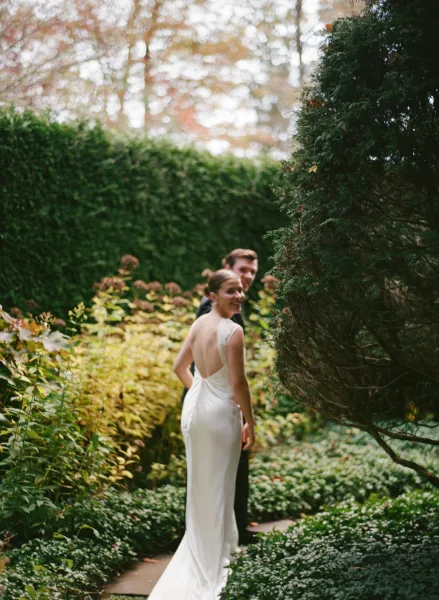 Couple portrait of bride and groom walking down a garden path, bride looking back in a backless wedding dress beside groom in suit
