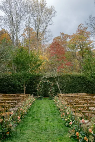 Outdoor ceremony setup with garden wedding ceremony aisle florals, wooden crossback chairs, and round rose arch on an overcast lawn with autumn trees