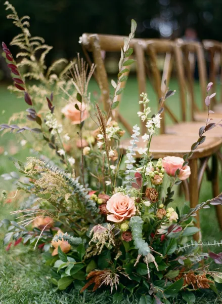 Ceremony aisle flowers with peach roses and eucalyptus accents lining wooden chairs on a grassy lawn, with trees behind the setup