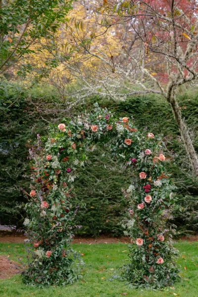 Floral wedding arch with peach and pink roses and greenery garland on a circular frame, set on a lawn with autumn trees behind