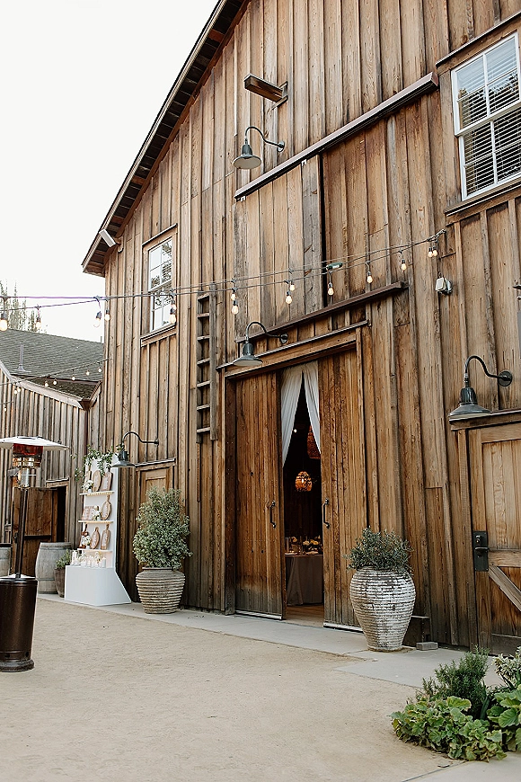 Barn wedding venue entrance with rustic barn exterior, open doors, string lights, escort card display, potted plants, and patio heater on concrete patio