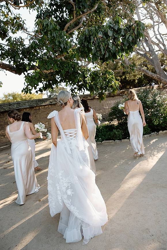 Bridesmaids walking from behind in satin white dresses holding bouquets, low bun hairstyles, along a sunlit garden pathway by a stone wall