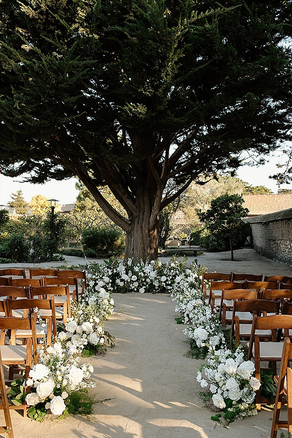 Ceremony aisle decor with outdoor wedding aisle flowers, white rose clusters and greenery garlands lining wooden chairs in a courtyard under a large tree