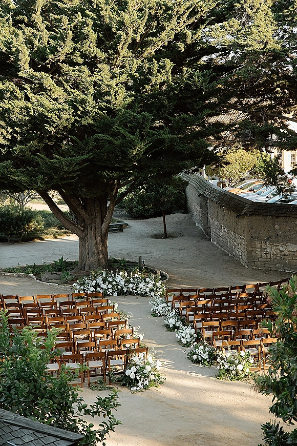 Outdoor ceremony setup with garden ceremony seating, wood folding chairs around a curved aisle lined with white florals and greenery under an evergreen tree