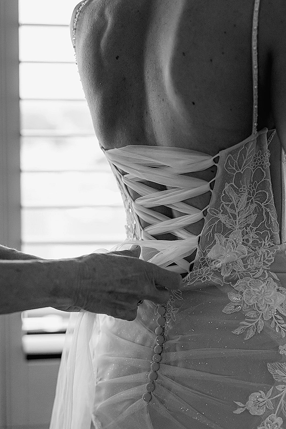 Wedding dress close-up showing corset back wedding dress lacing, lace appliques and bridal buttons in soft window light through blinds