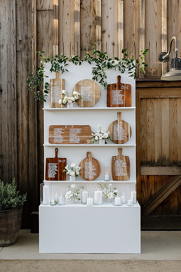Seating chart display with a wedding seating chart on white shelves, wooden cutting boards, greenery garland, and candles against a barn wall